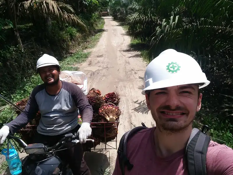 Luca Fischer in a hard hat on a plantation road, next to a worker carrying fresh palm fruit bunches on a motorbike